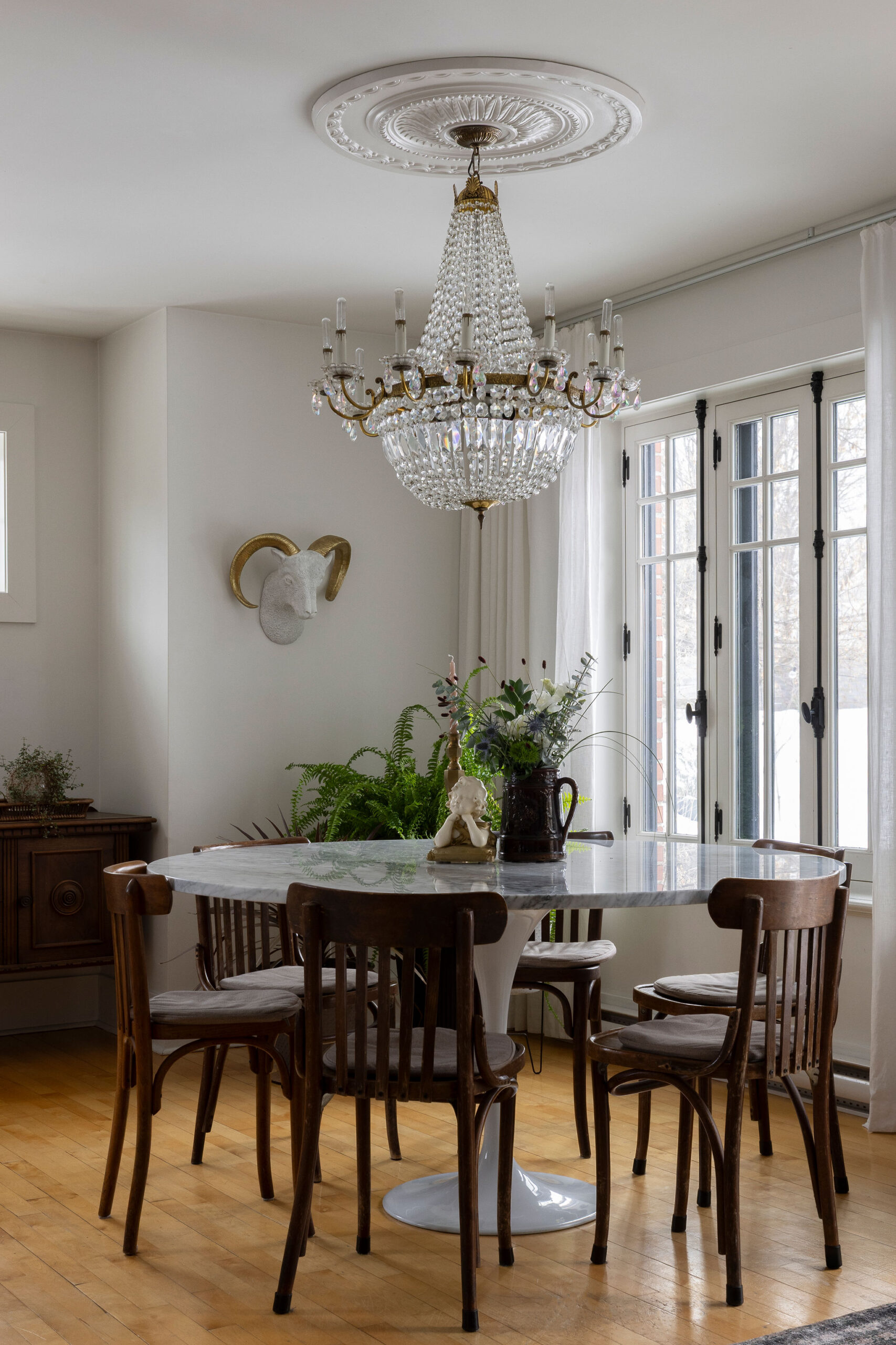 Salle à manger lumineuse avec table ronde blanche, chaises en bois, lustre antique en cristal, grandes portes vitrées et bouquet de fleurs.