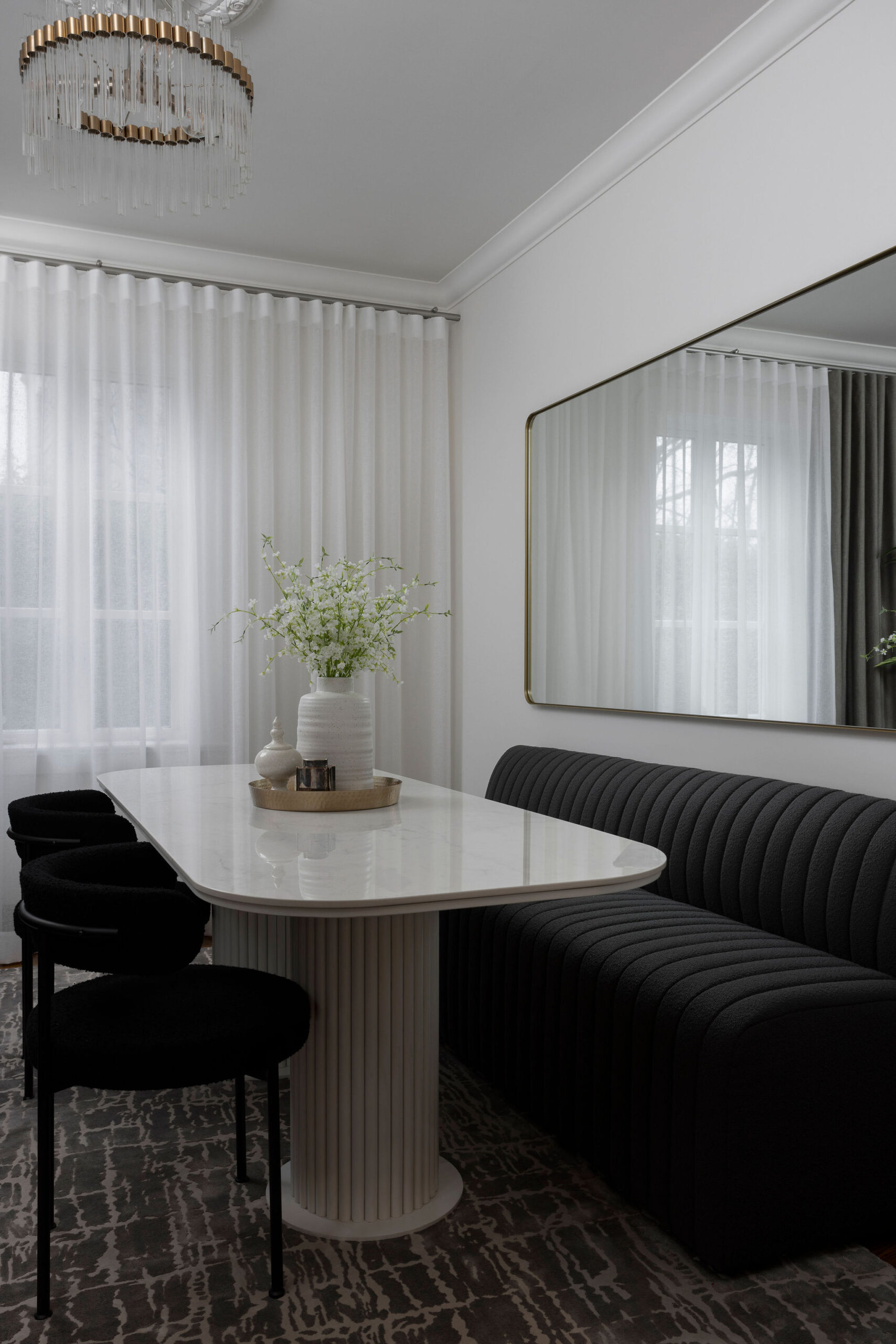 Coin repas lumineux avec table ronde blanche, banquette foncée, chaises noires, voilages blancs et moulures murales.