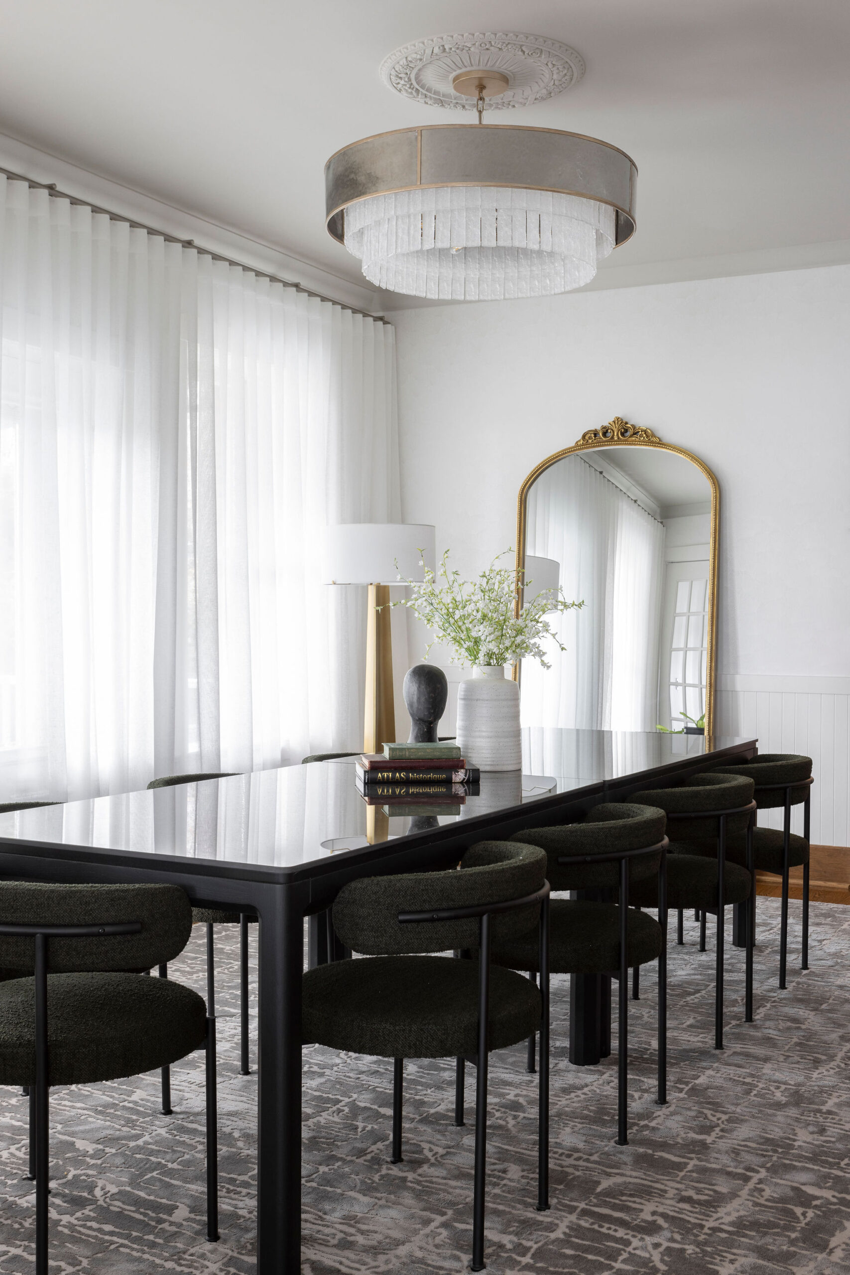Salle à manger lumineuse avec longue table noire, chaises noires, voilages blancs, grand miroir arqué et sol à motif.