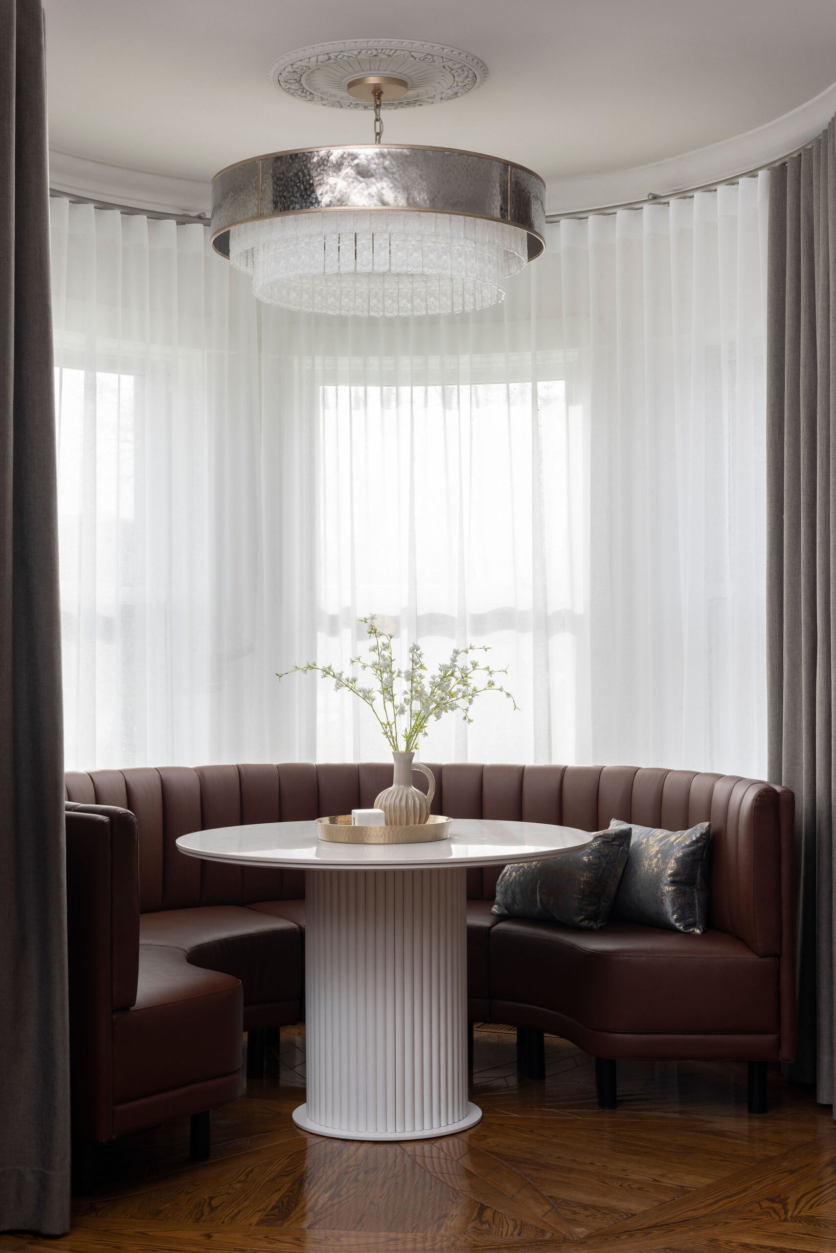 Coin repas avec table ronde blanche, assises foncées, grande fenêtre voilée, vase sur la table, banquette demi-lune et ambiance douce.